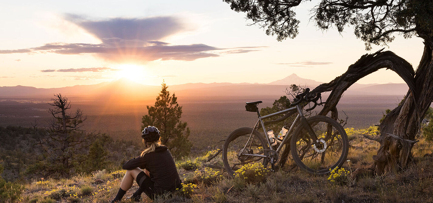 Shimano GRX Radfahrerin sitzt im Abendlicht auf einer Anhöhe und blickt ins Tal, an einem Baum lehnt ihr Gravelbike.